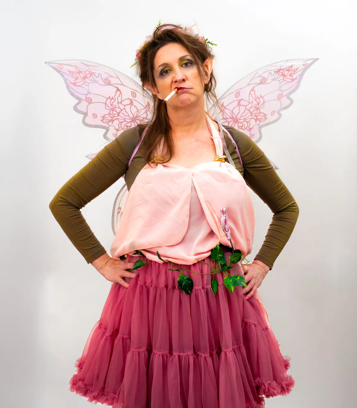 Rama Nicholas as Thistle, a scowling fairy with pink wings, pointed ears, a flower crown and a cigarette, hands on hips.