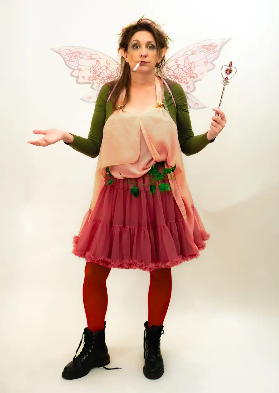 Thistle shrugging with her wand in hand, wearing a pink tutu and red tights against a white studio wall.